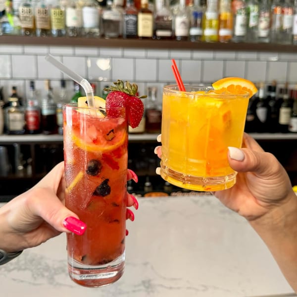 Two hands holding up a red fruit cocktail and an orange cocktail with a bar in the background.