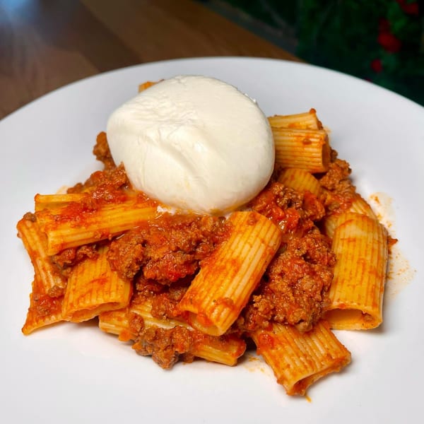 Rigatoni pasta with rich bolognese sauce and a large dollop of fresh burrata cheese on a white plate.