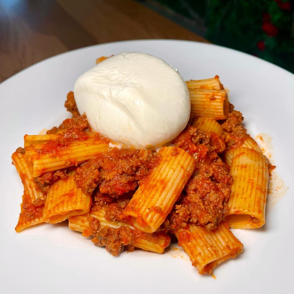 Rigatoni pasta with rich bolognese sauce and a large dollop of fresh burrata cheese on a white plate.