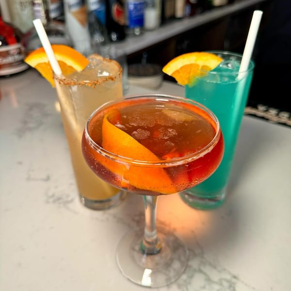 Three colorful cocktails, one with an orange peel, another with an orange slice, on a white bar.