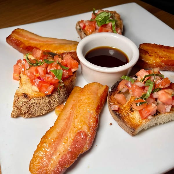 A white plate with bruschetta, thick-cut bacon, and a small bowl of dark sauce.