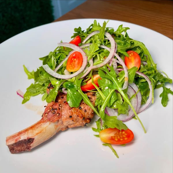 Grilled lamb chop with a fresh arugula salad, cherry tomatoes, and red onion on a white plate.
